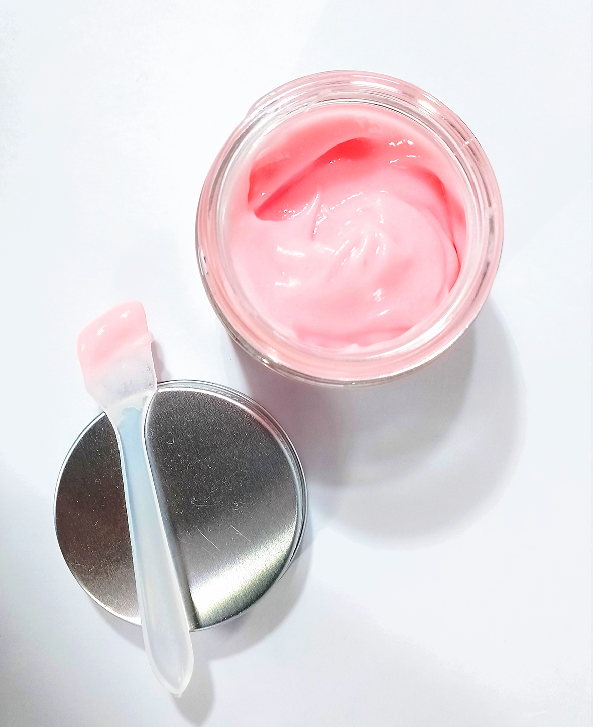 Telový jogurt ružovej farby v okrúhle nádobke s priehľadnou plastovou naberačkou vo formy lyžičky leží na bielom pozadí.